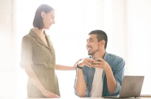 man sitting at table with woman standing and putting her hand on his shoulder, both are smiling